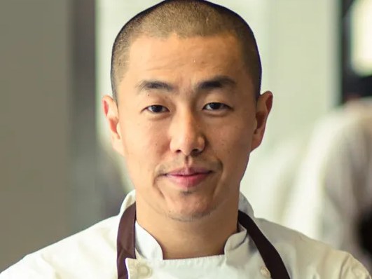 Portrait of an Asian male chef in white chef's coat and dark apron standing in a professional kitchen; source page explicitly identifies this as Corey