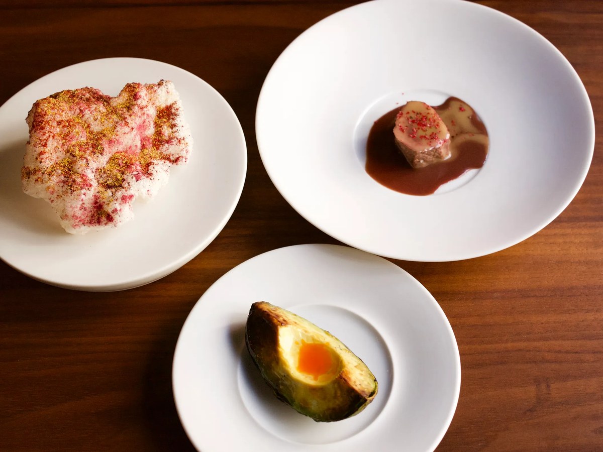 Three white plates on a wooden table, each containing a single carefully plated dish: a pink-dusted dessert, a seared protein with sauce, and a roasted vegetable with a bright center.
