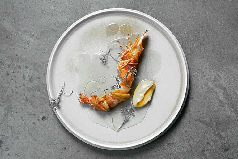 A plated fine-dining dish featuring a tiger prawn with lemon wedge on a ceramic plate.