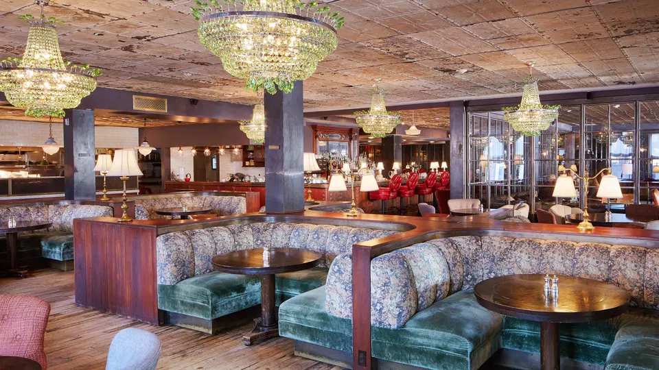 An empty dining room at Soho House New York with green velvet banquettes, round tables, crystal chandeliers, and a bar in the background.