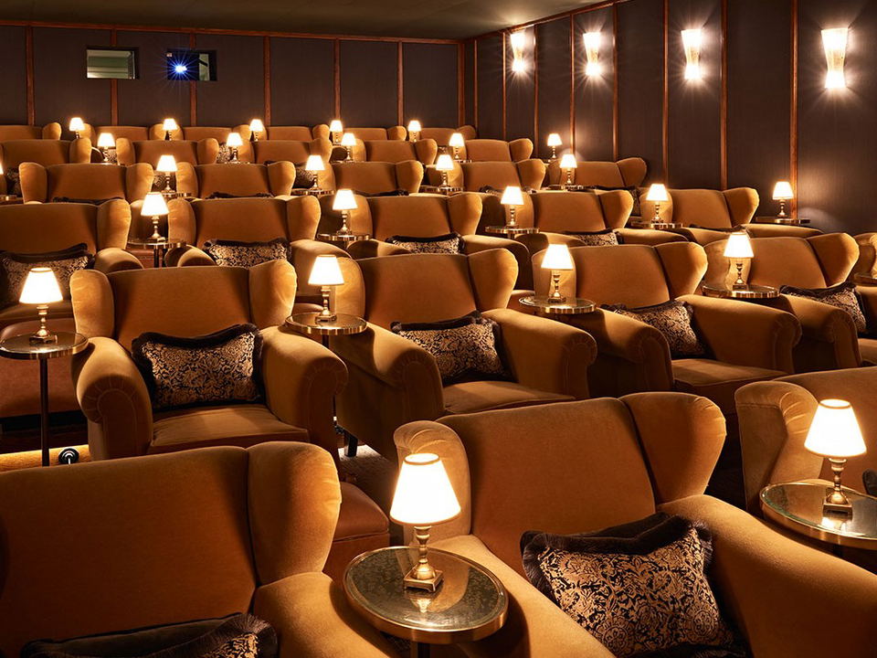 A luxurious private cinema screening room with rows of plush caramel-colored velvet armchairs, small side tables with lamps, and wall sconces.