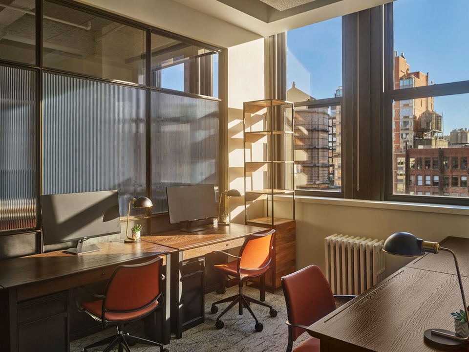A sunlit office workspace with dark wood desks, orange chairs, and a brass-framed shelving unit, overlooking a New York City skyline.