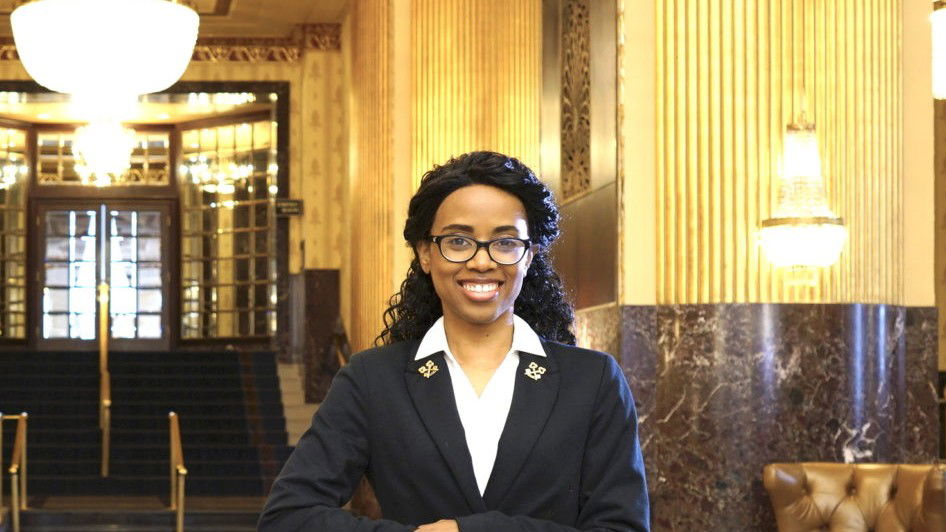 A smiling female concierge in a dark blazer stands behind a glass-topped desk in a grand hotel lobby.