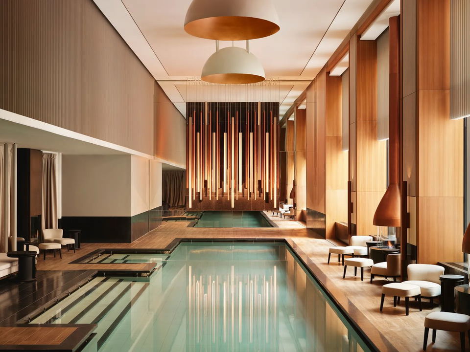 A luxurious indoor pool and spa area with warm wood paneling, pendant lighting, lounge seating with fireplaces, and a dramatic illuminated
