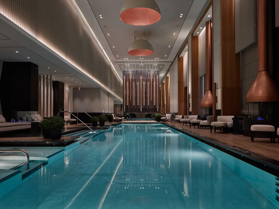 A luxurious indoor pool at Aman New York with turquoise water, warm wood paneling, elegant lounge seating, and suspended fireplaces.