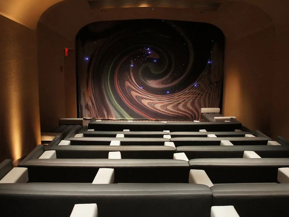 A luxury private screening room with tiered rows of dark leather loungers facing a decorative swirling-galaxy curtain/screen.