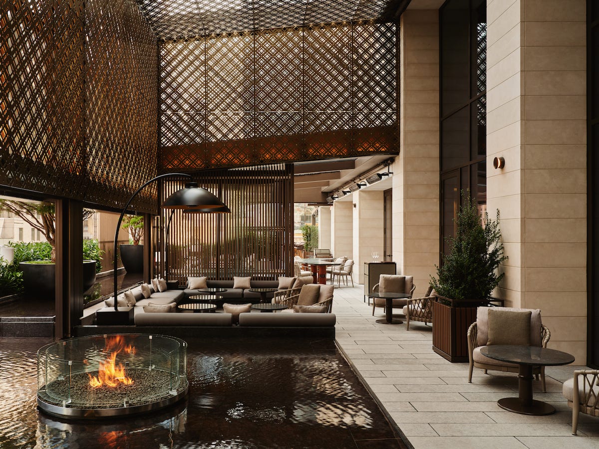 A luxurious indoor courtyard with a circular fire pit, beige upholstered seating, black metal architectural elements, potted plants, and geometric patterned ceiling screens casting shadows on the floor.