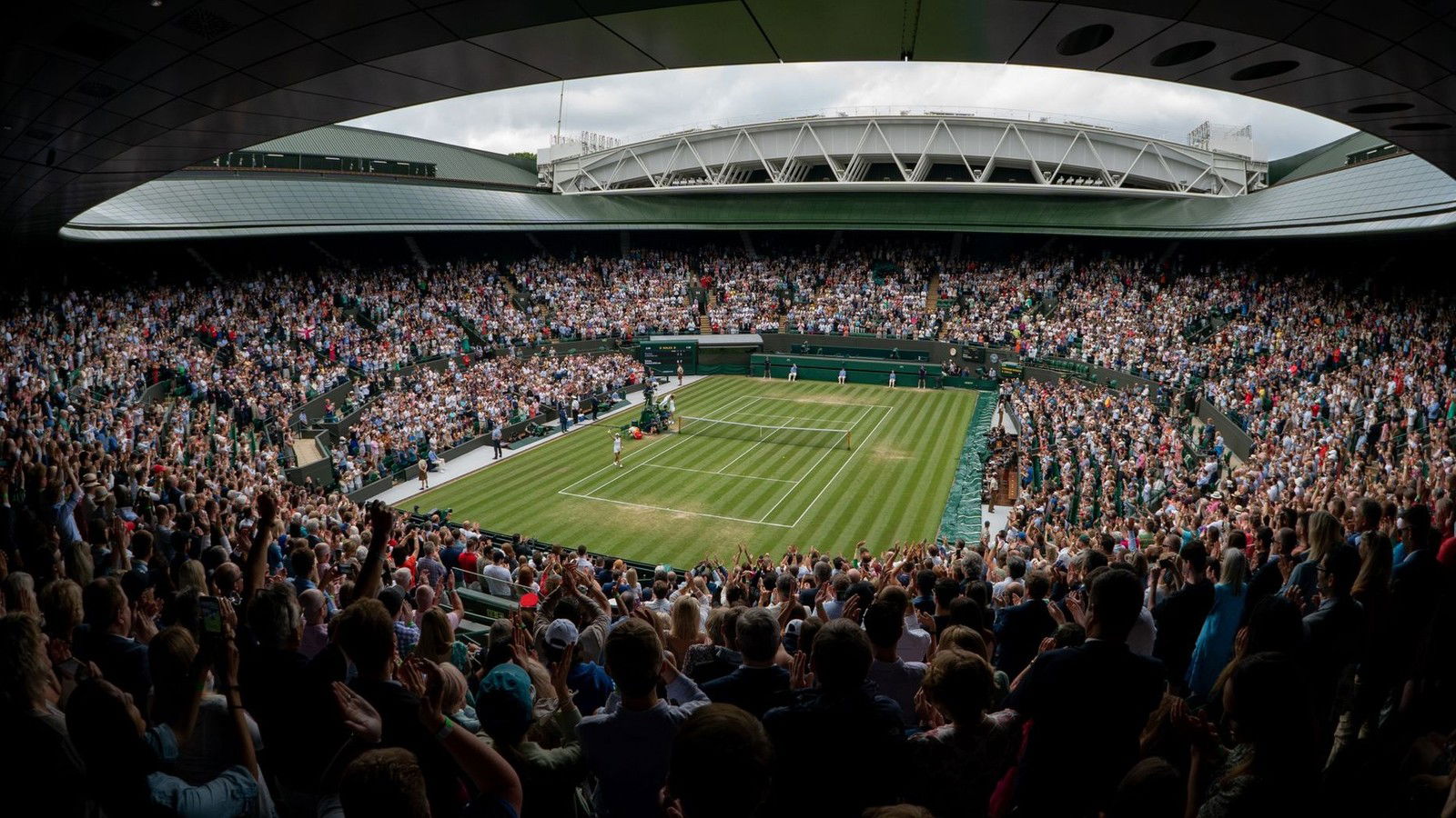 A wide-angle panoramic view of Centre Court at the All England Club, showing the retractable roof, packed stands, and a tennis match in progress.