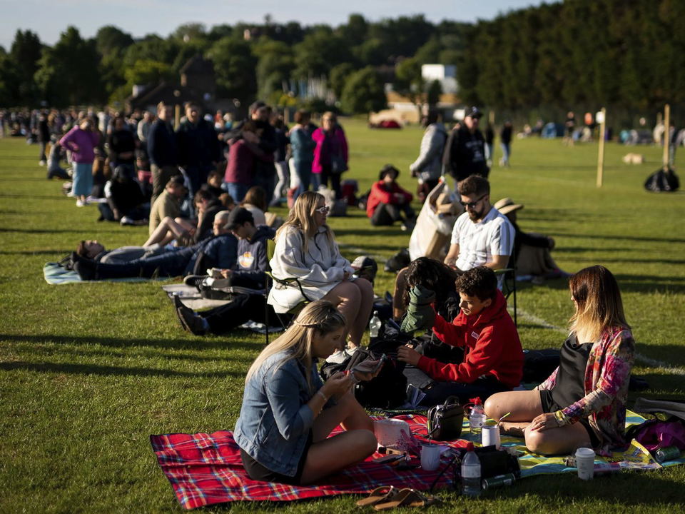 A large crowd of people are sitting and lying on a grassy field under a clear sky, enjoying a sunny day.