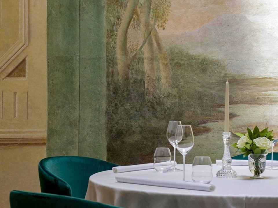 An elegantly set dining table with white linen, crystal glasses, a taper candle, and white roses, against a classical landscape fresco.