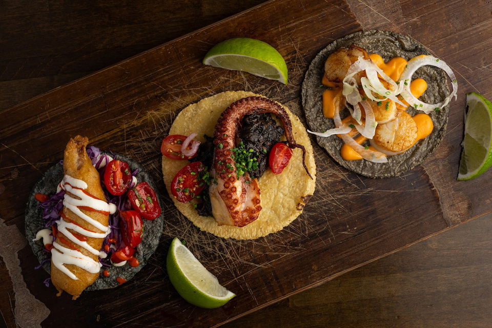Holbox tacos topped with octopus, fish, vegetables, and lime wedges on a wooden table.
