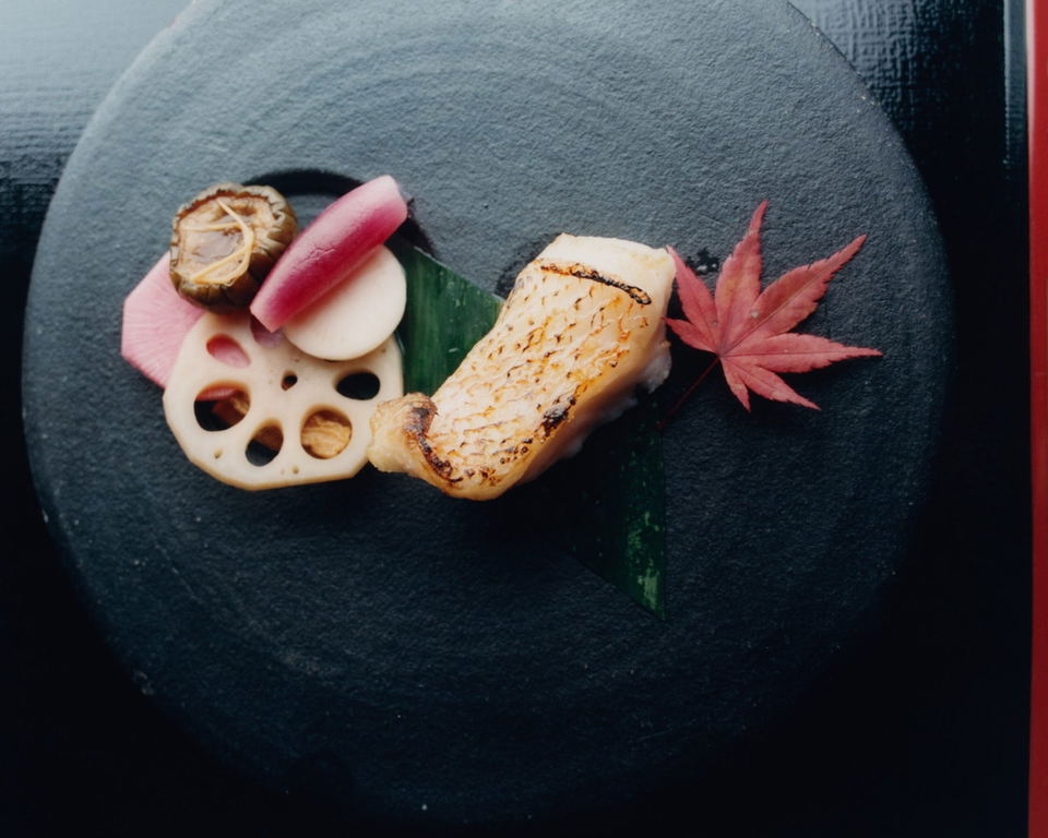Morihiro dish with grilled fish, lotus root, radish, mushroom, and a red maple leaf on a black plate.