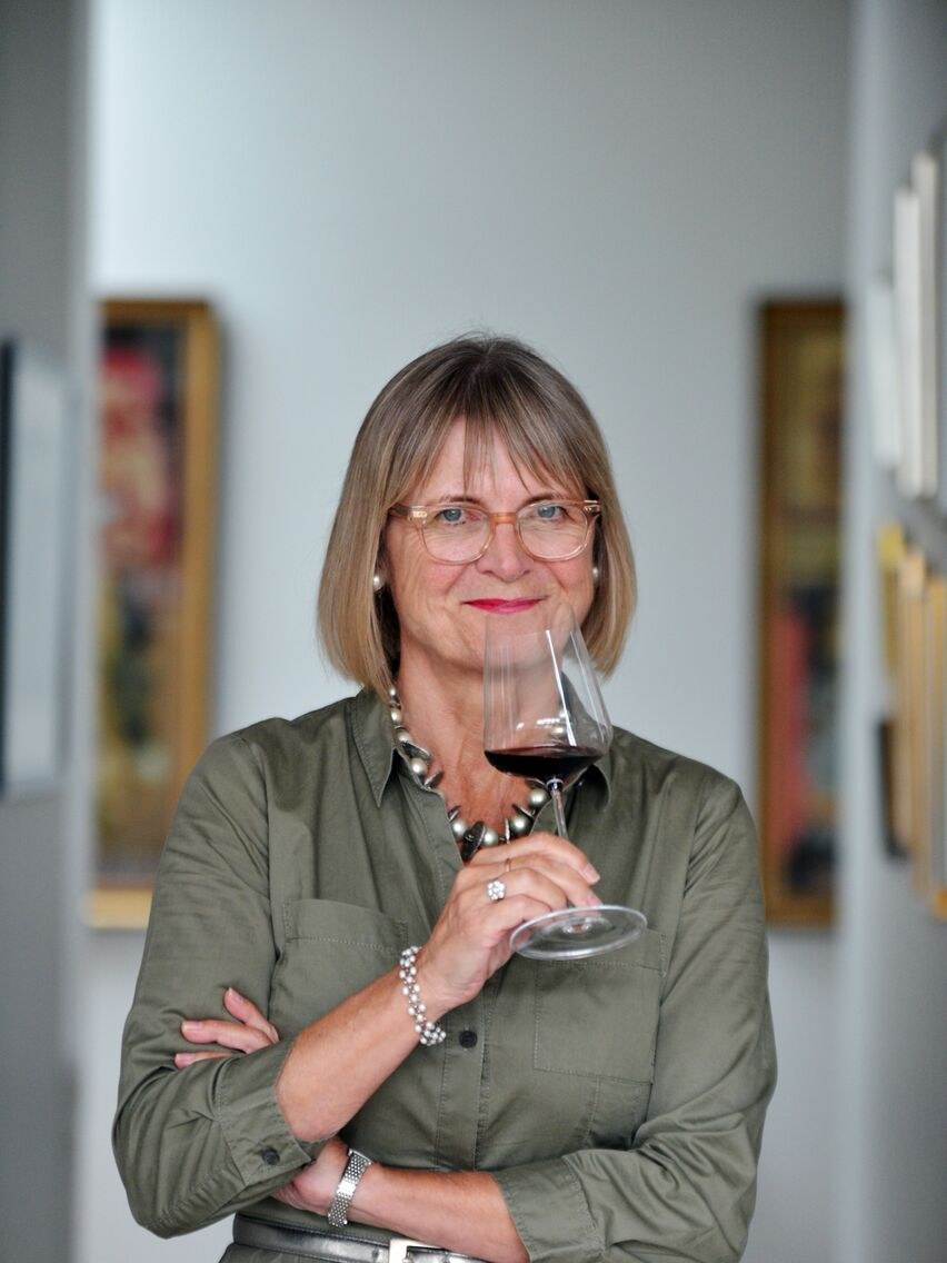Professional portrait of a woman with bob haircut and rose-tinted glasses holding a wine glass in an art gallery setting