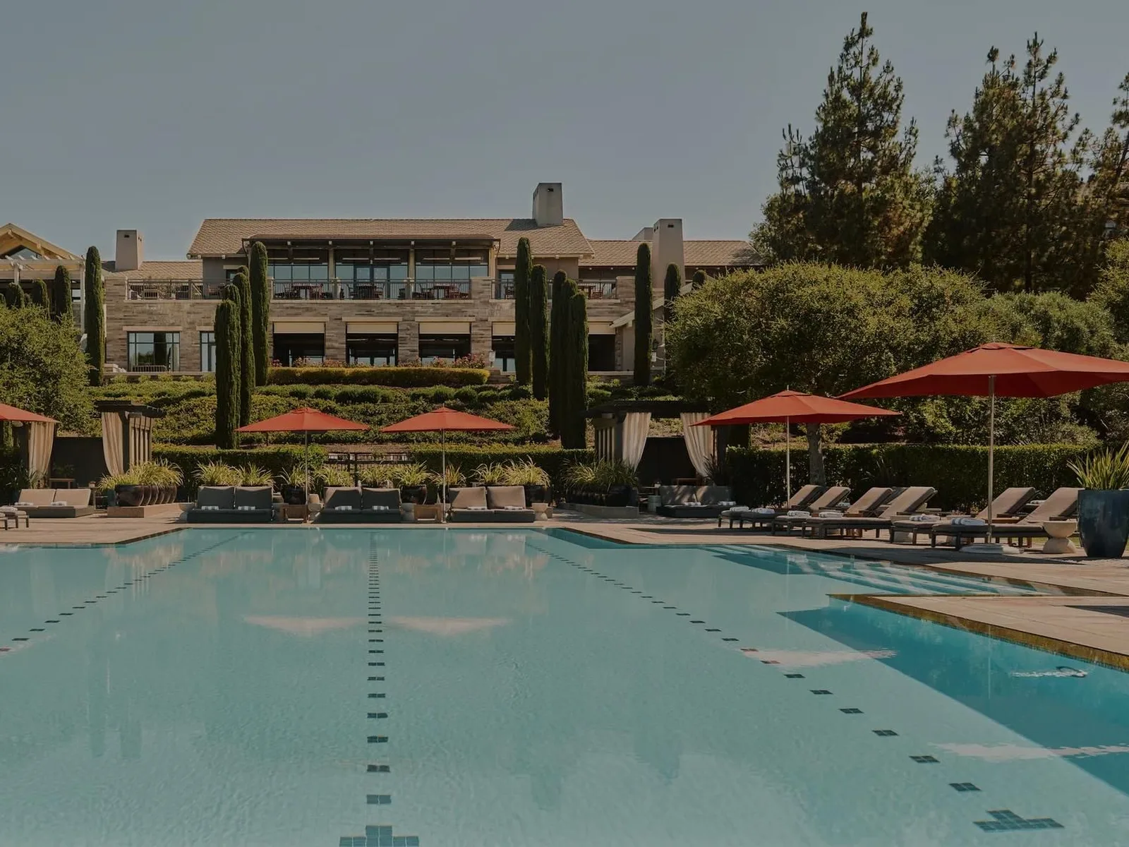 A large, multi-story stone building with a tiled roof, surrounded by lush green landscaping and tall trees, overlooks a swimming pool with red