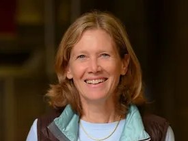 Portrait of a smiling woman with reddish-blonde hair wearing a light blue scarf over dark clothing.