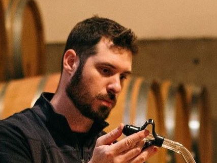 A winemaker with a beard draws a sample of red wine from a barrel using a wine thief in a cellar filled with oak barrels.