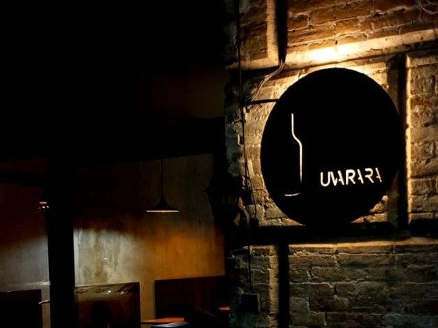 A dimly lit wine bar interior with exposed brick walls, dark wood booth seating, and a circular black sign with a wine bottle silhouette and the word