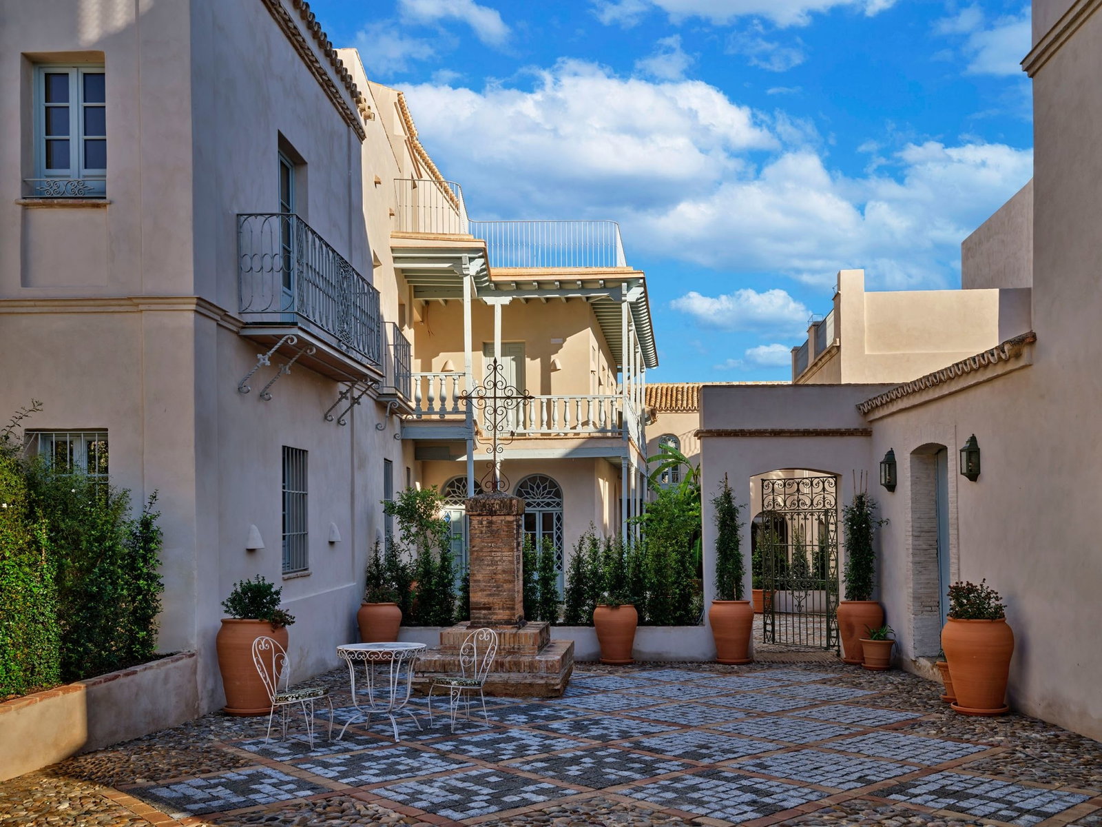 A courtyard view of a historic Spanish estate with Mediterranean architecture, terracotta planters, wrought-iron details, balconies, and cobblestone p