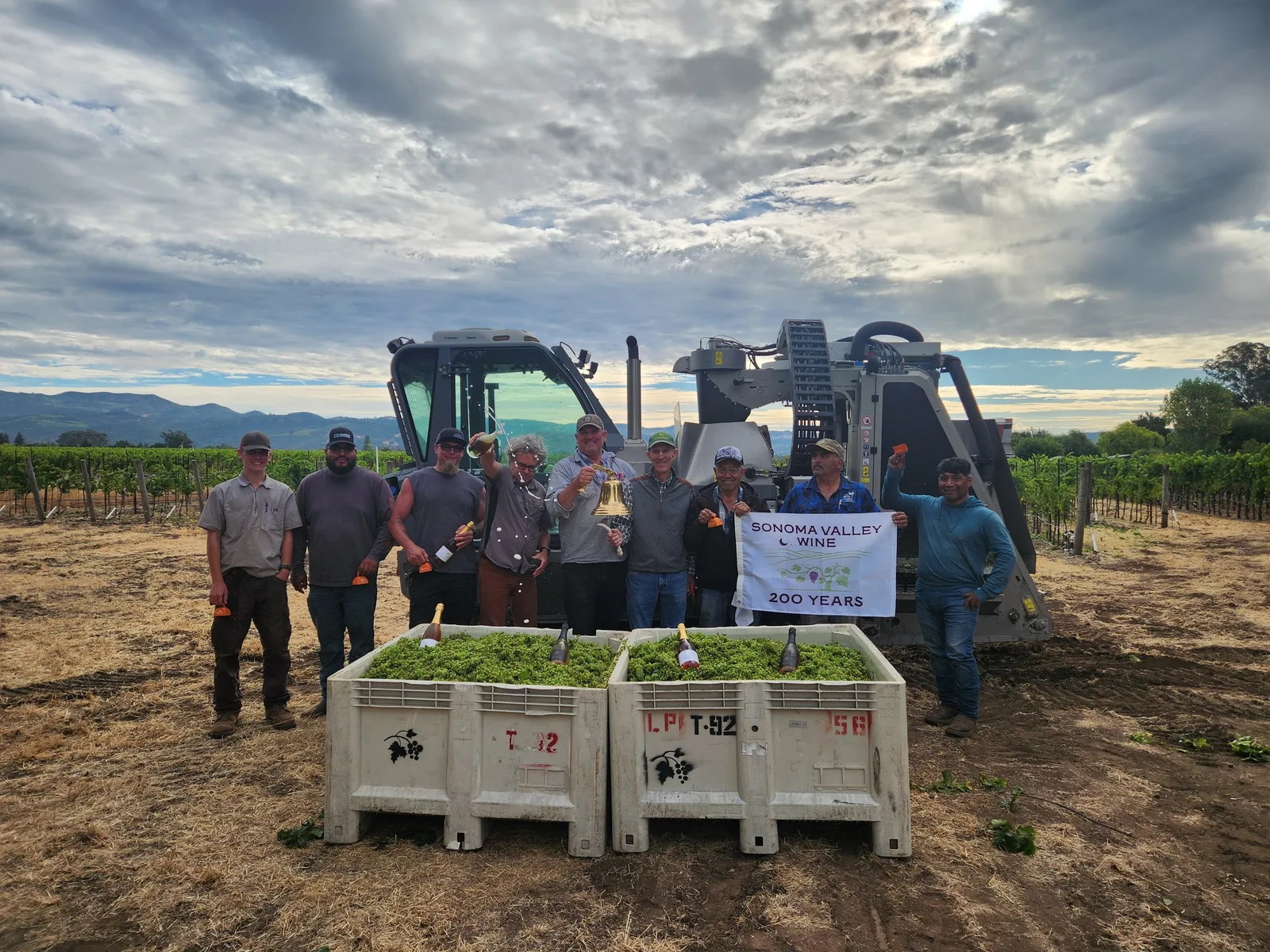 Ten men in a vineyard with a tractor, two large bins of grapes, and a banner reading "Sonoma Valley Wine 200 Years."