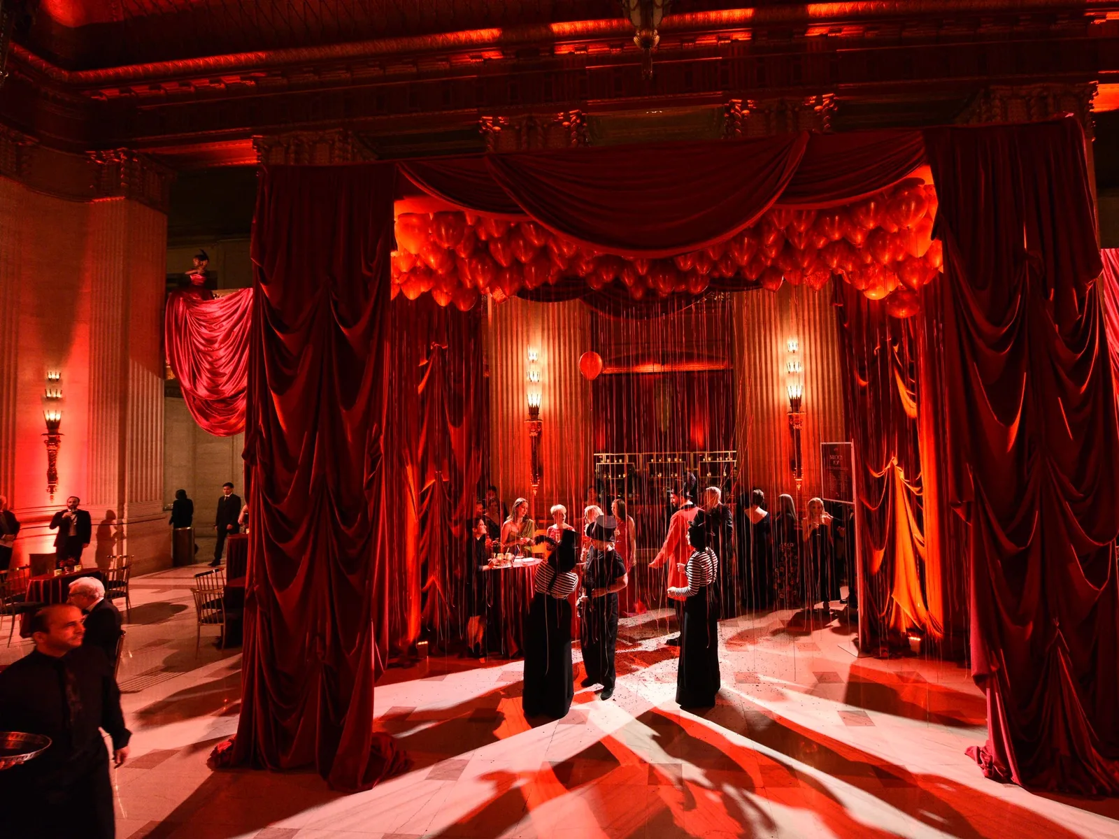 A grand hall decorated with red drapes and balloons, illuminated by red lighting, with guests mingling at an event.