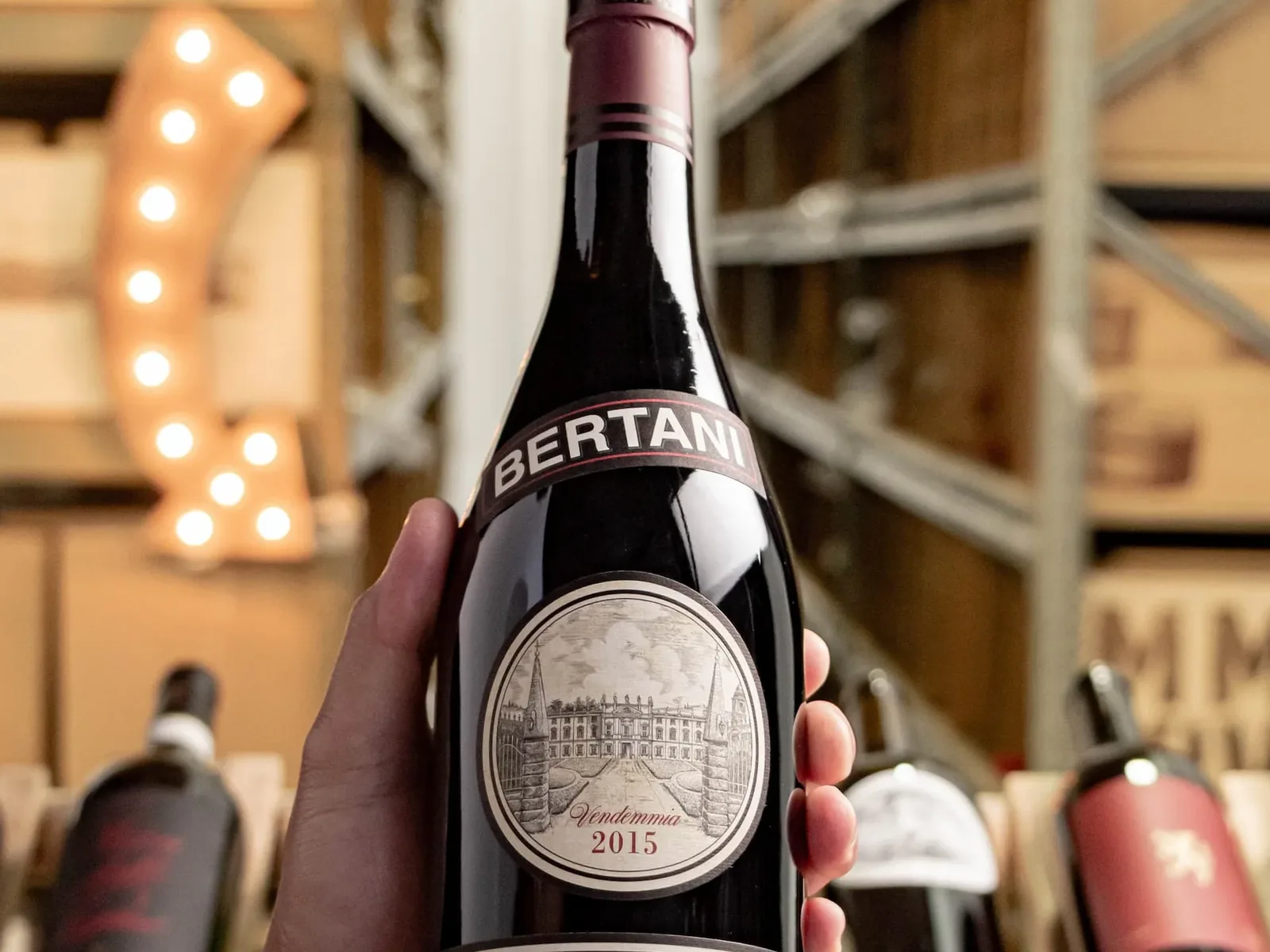 A hand holds a bottle of Bertani Amarone della Valpolicella Classico 2015, with other wine bottles blurred in the background.