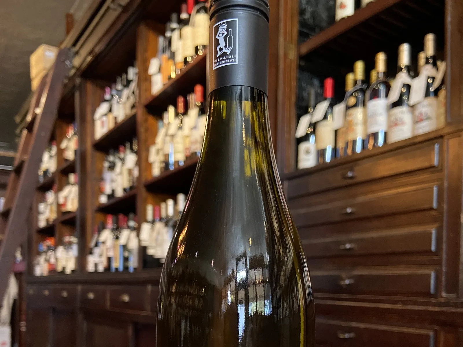 A bottle of Claudio Mariotto Derthona white wine from 2023, with a black and white label, against a backdrop of wine shelves.