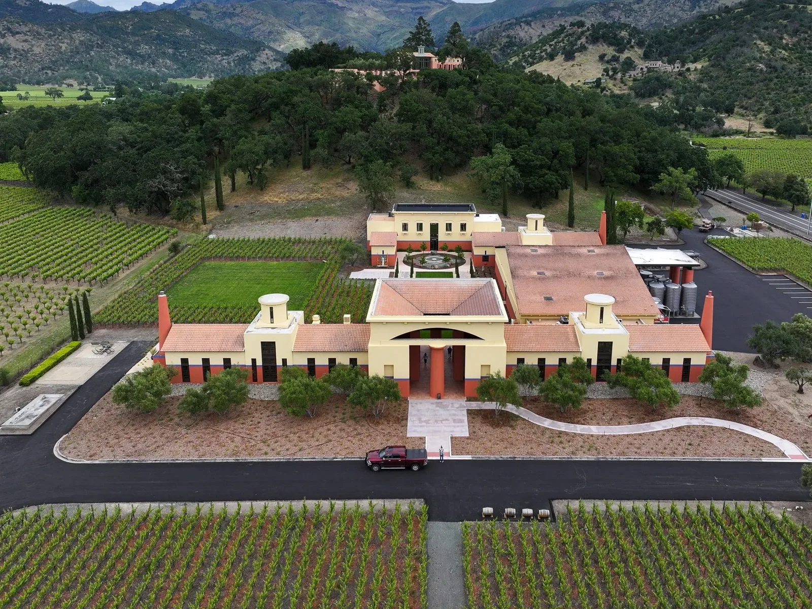 An aerial view of Clos Pegase Winery, showing its distinctive architecture, surrounding vineyards, and distant mountains under a cloudy sky.