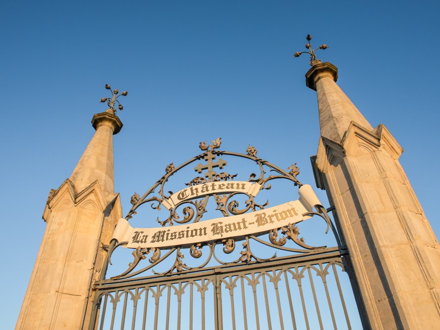 An ornamental iron gate with decorative scrollwork and text reading 'Chateau La Mission Haut-Brion' mounted between two stone pillars topped with crosses against a blue sky.
