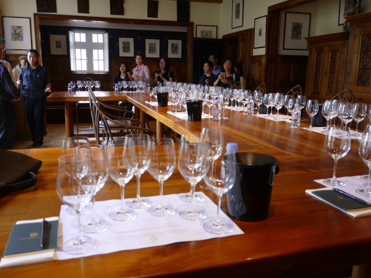 A wine tasting session in progress in an elegant wood-paneled room with numerous wine glasses arranged on tables and guests sampling wines; the source