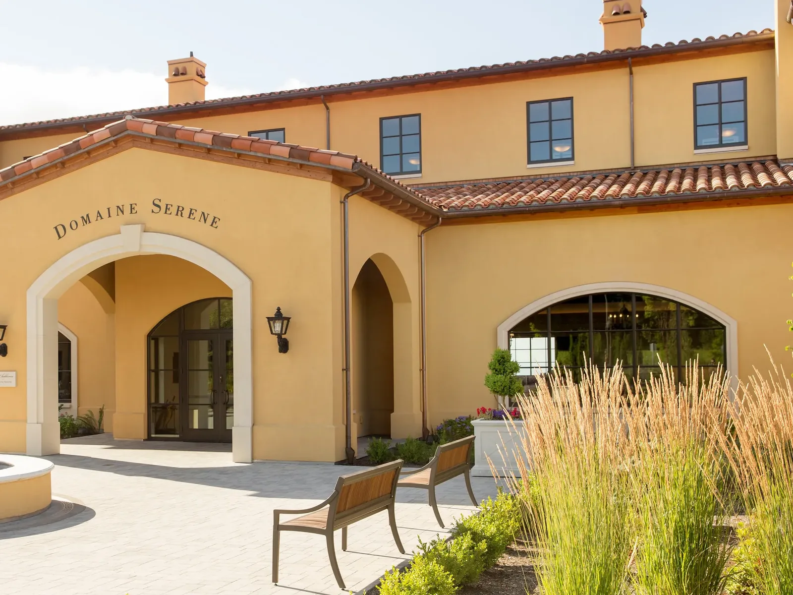 A yellow stucco winery building with white-trimmed arched doorways, terracotta roof tiles, decorative chimneys, and ornamental grasses in the foreground.