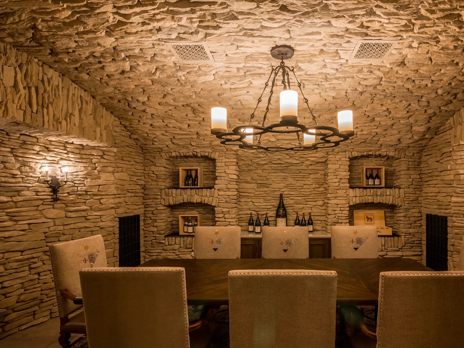Interior of a stone-walled underground wine cellar with a central chandelier, three service counters, and built-in shelving displaying wine bottles.