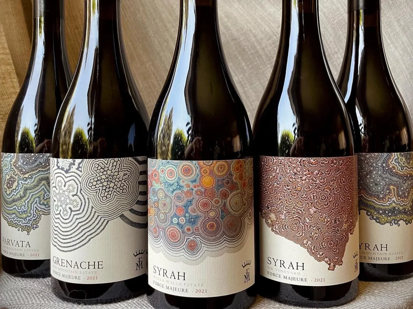 Five wine bottles from Force Majeure Estate arranged in a row, showing dark wine inside glass bottles with colorful geometric and organic patterned labels, all dated 2021.