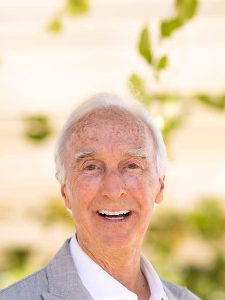 An elderly man with white hair smiling warmly at the camera, wearing a gray blazer and white shirt, photographed outdoors with blurred foliage in the background.