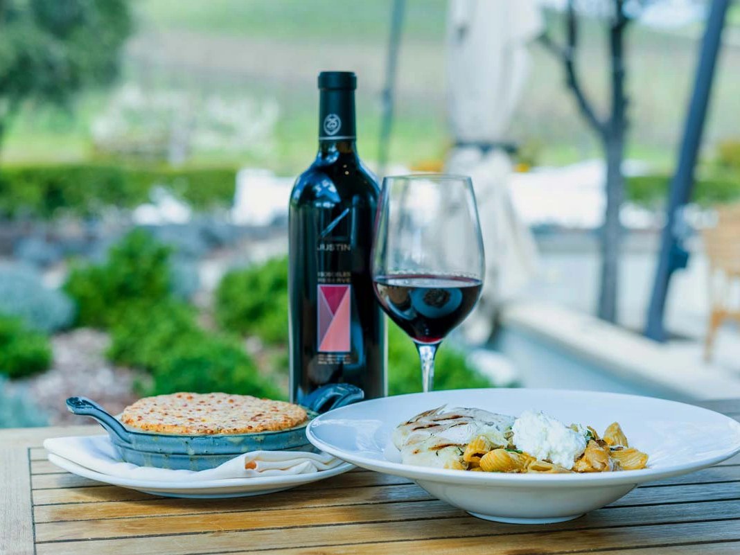 A wine bottle labeled JUSTIN with a glass of red wine and plated food on an outdoor table; the source page explicitly attests this is from JUSTIN Wine