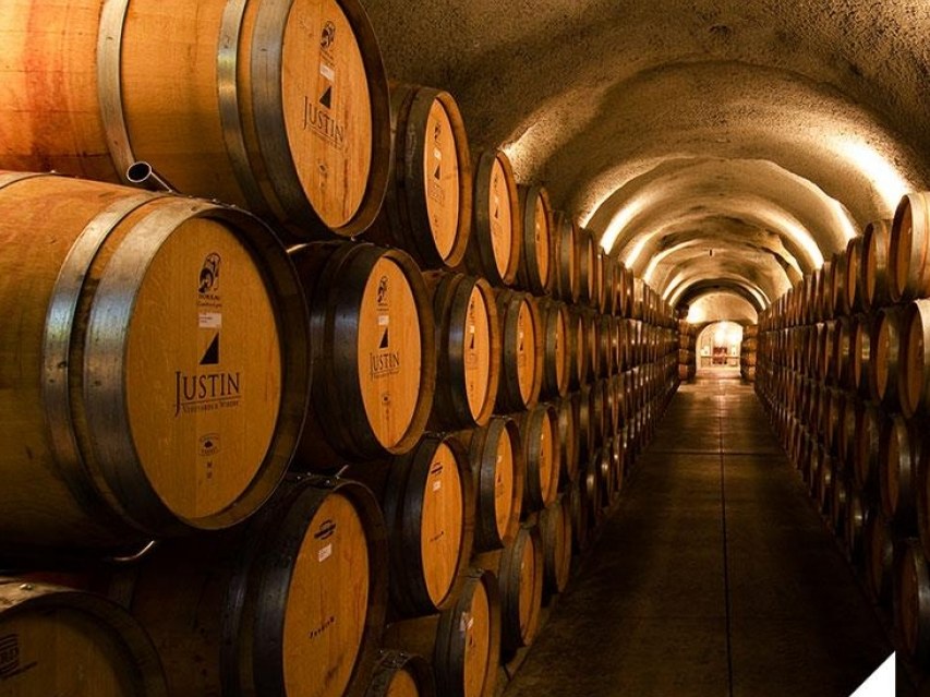 An underground wine cellar with arched stone ceiling and rows of oak barrels stacked along both sides; the source page explicitly attests this is JUST