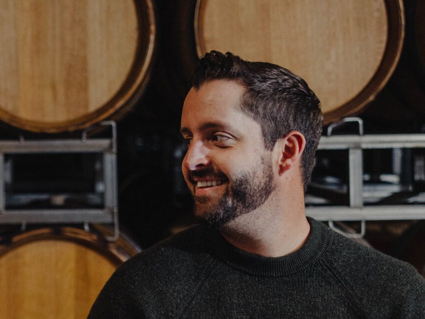 A bearded man in a dark sweater stands in front of large wooden barrels stacked on metal racks in a winery cellar.