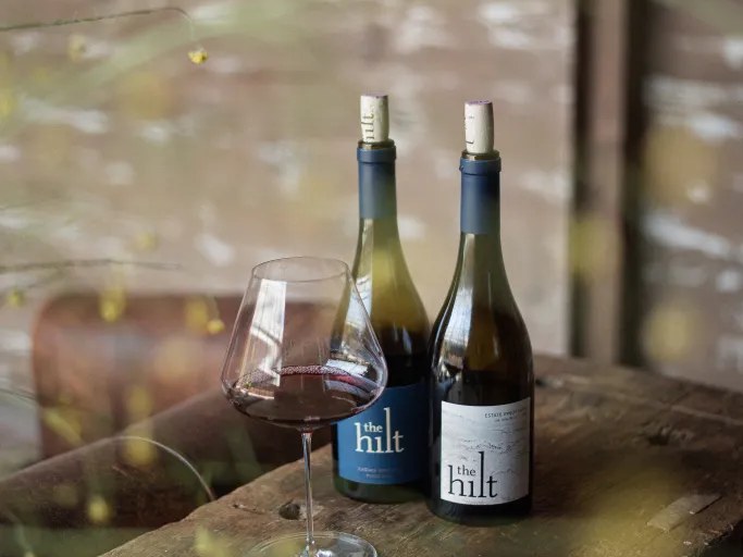 The Hilt offers a taste of their finest, with two bottles and a glass of red wine on a rustic table.