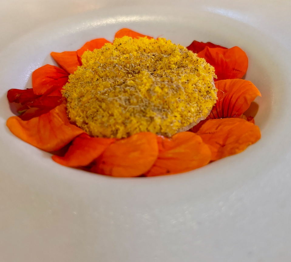 A round yellow crumb-coated portion rests in a white bowl, surrounded by bright orange and red edible flower petals.
