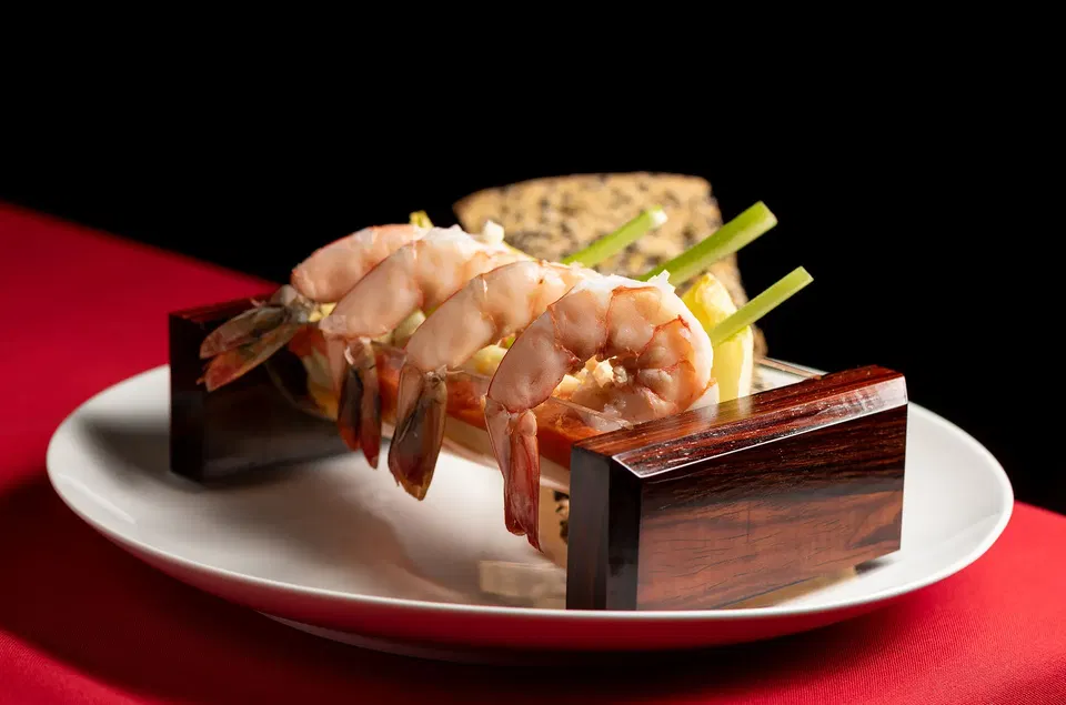Large poached shrimp draped over polished dark wood blocks on a white plate, with celery sticks and a textured cracker, on a red tablecloth.