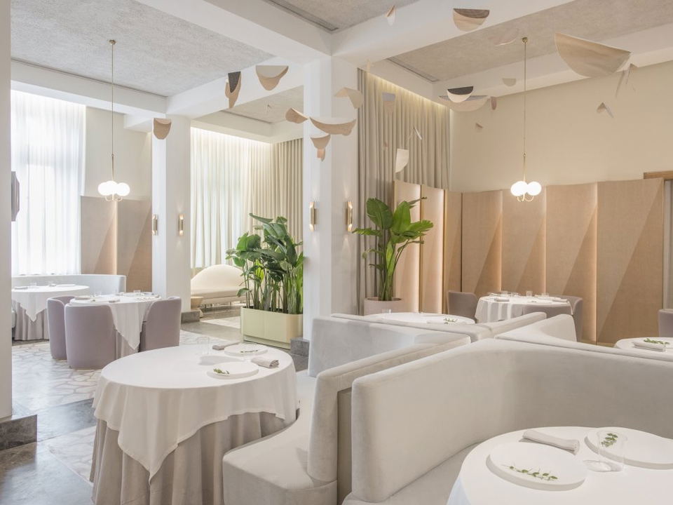 An elegant, minimalist fine-dining interior with a neutral palette, sculptural ceiling mobiles, velvet upholstered seating, and tropical plants.