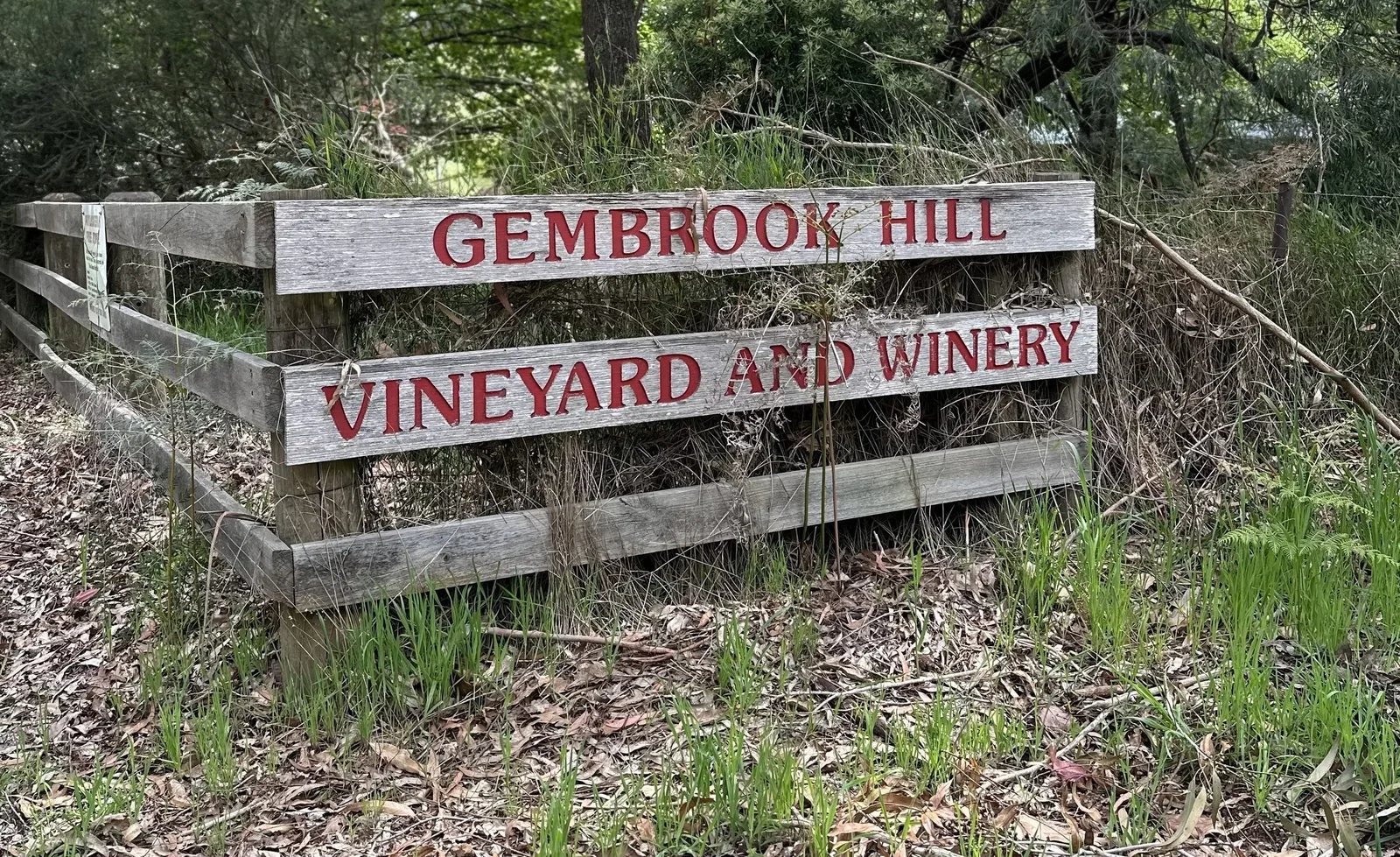 Wooden fence sign reading 'Gembrook Hill Vineyard and Winery' surrounded by native vegetation and mulch