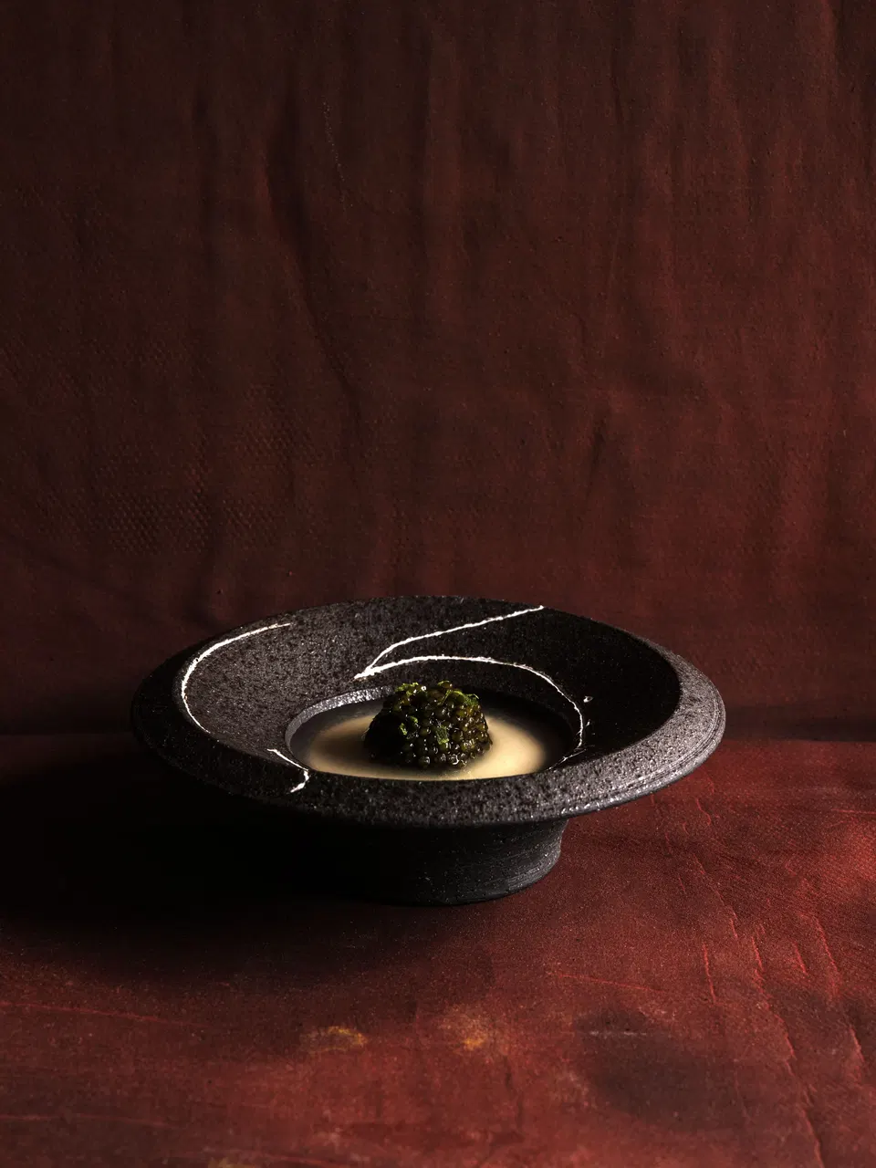 A close-up of a custom black pottery bowl with white decorative lines containing a plated dish with caviar on a cream sauce, photographed on a texture