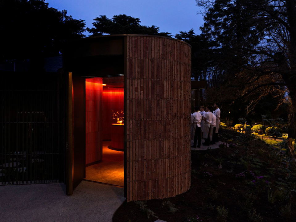 A modern wooden pavilion structure photographed at dusk in a garden setting with warm interior lighting