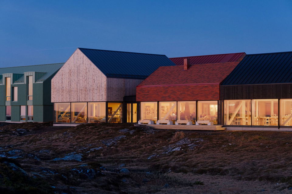 Contemporary architecture featuring multiple connected structures with colored metal roofing and extensive windows, illuminated from within, set against a barren terrain under a blue twilight sky.