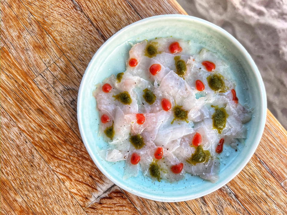 A light blue ceramic bowl containing translucent sliced raw fish (likely a crudo or ceviche preparation) garnished with red dots (possibly chili oil o