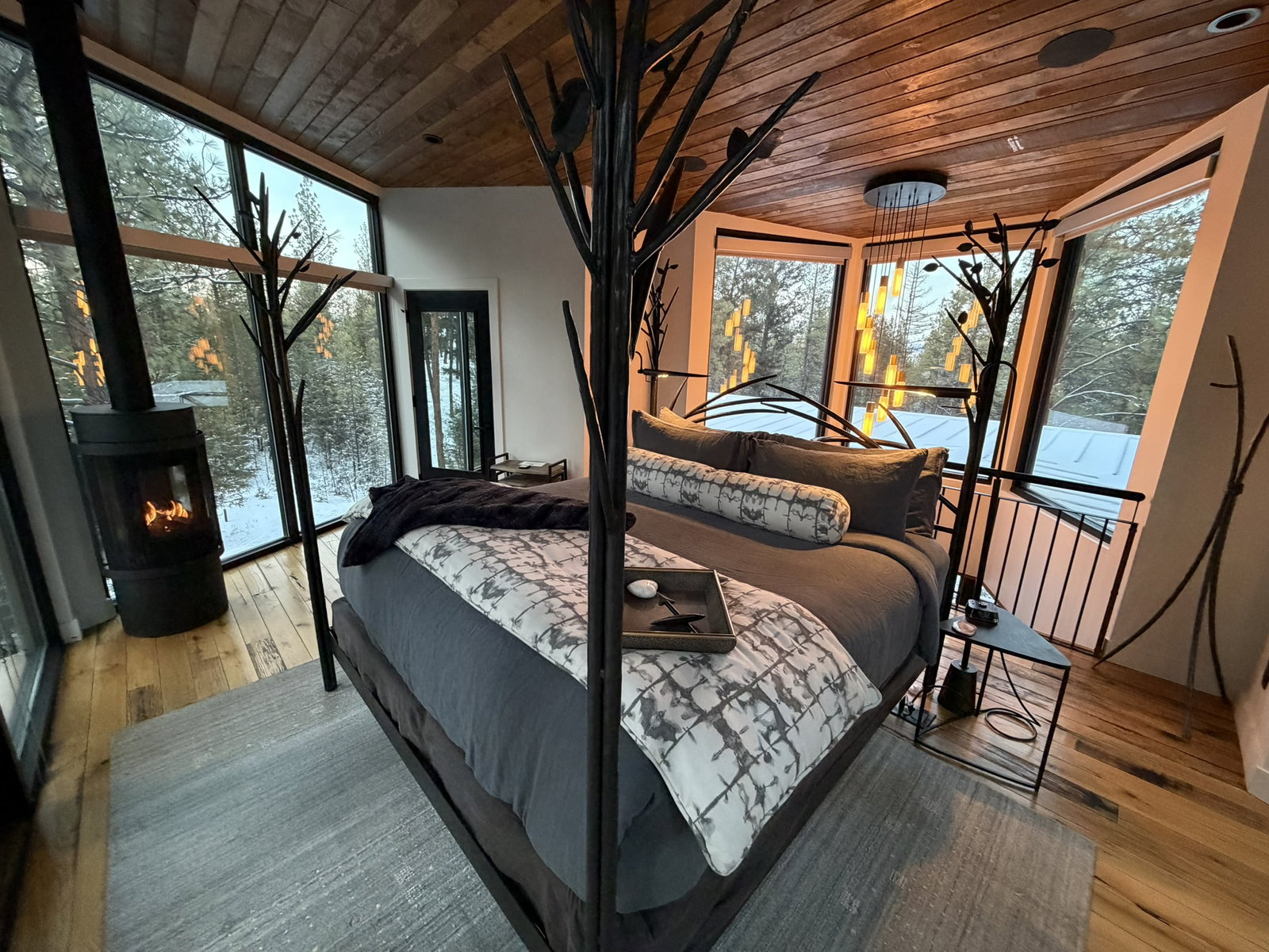 Cozy bedroom with large windows showing snowy forest, a lit fireplace, tree branch decor, patterned bedding, and warm lighting, creating a serene ambiance.