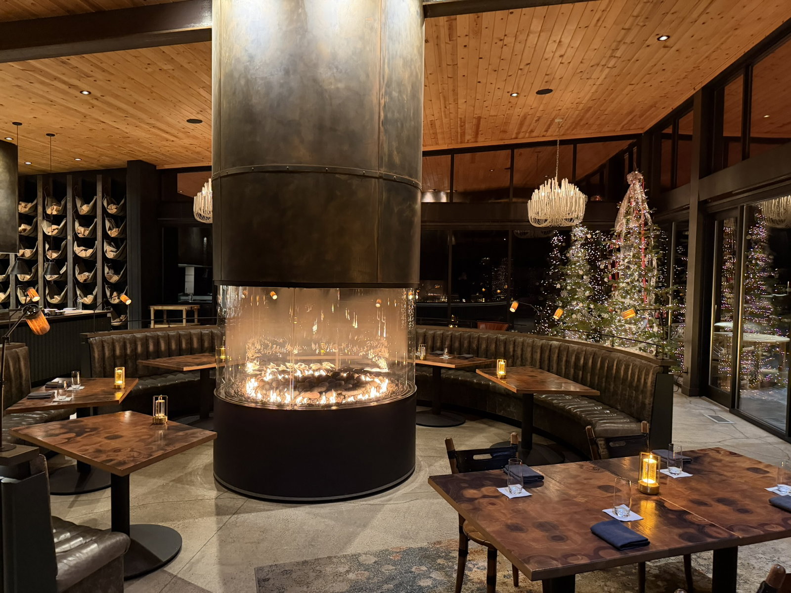 Cozy restaurant with a central modern fireplace, wooden tables, and leather seating. Festive trees and warm lighting enhance the elegant mood.