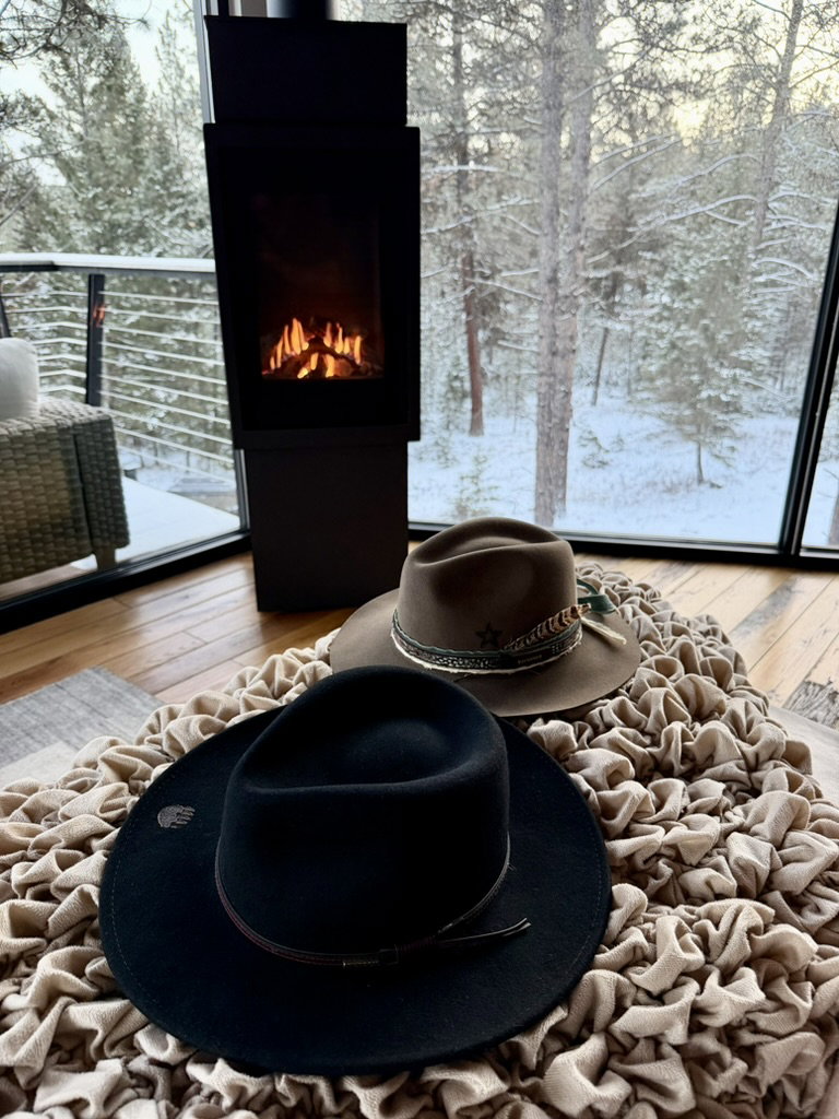 Two hats on a textured beige cushion by a fireplace, with snow-covered trees visible through the glass doors in the background. Cozy mood.