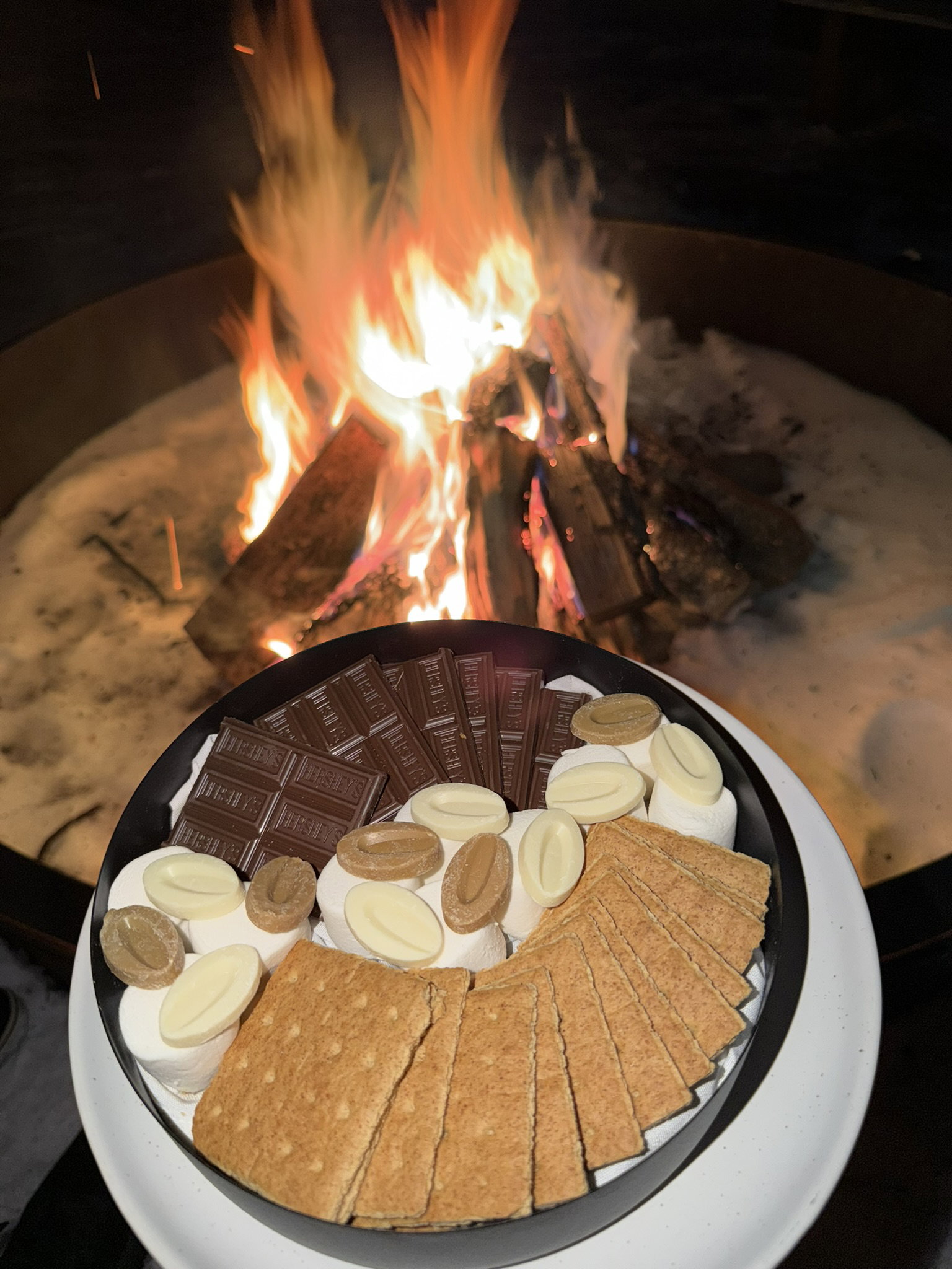 Plate with marshmallows, chocolate, and crackers near a blazing fire pit at night, creating a cozy atmosphere. Chocolate marked with Hershey's.