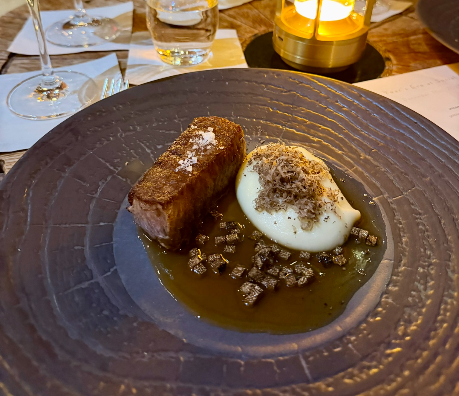Steak with mashed potatoes and truffle on a textured, dark plate. Warm candlelight in the background creates a cozy dining ambiance.